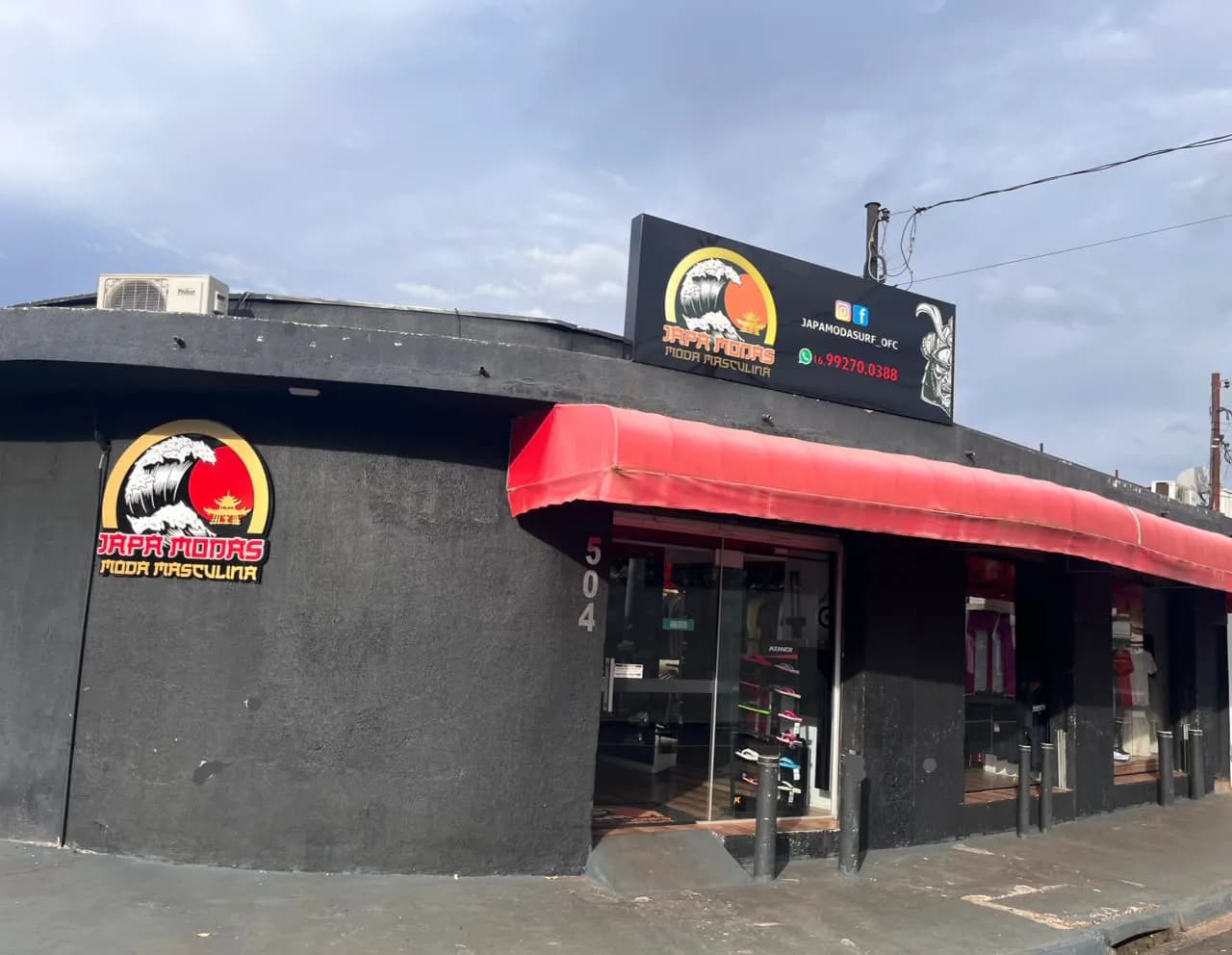 A fachada da Japa Modas: parede preta com a logo da onda, a placa, o número 504 na porta e o toldo vermelho